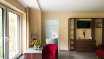 Hotelkamer met een groot raam, een bureau met een bloevas, een rode stoel, een bed, een dressoir, een spiegel en een televisie op een houten kast.