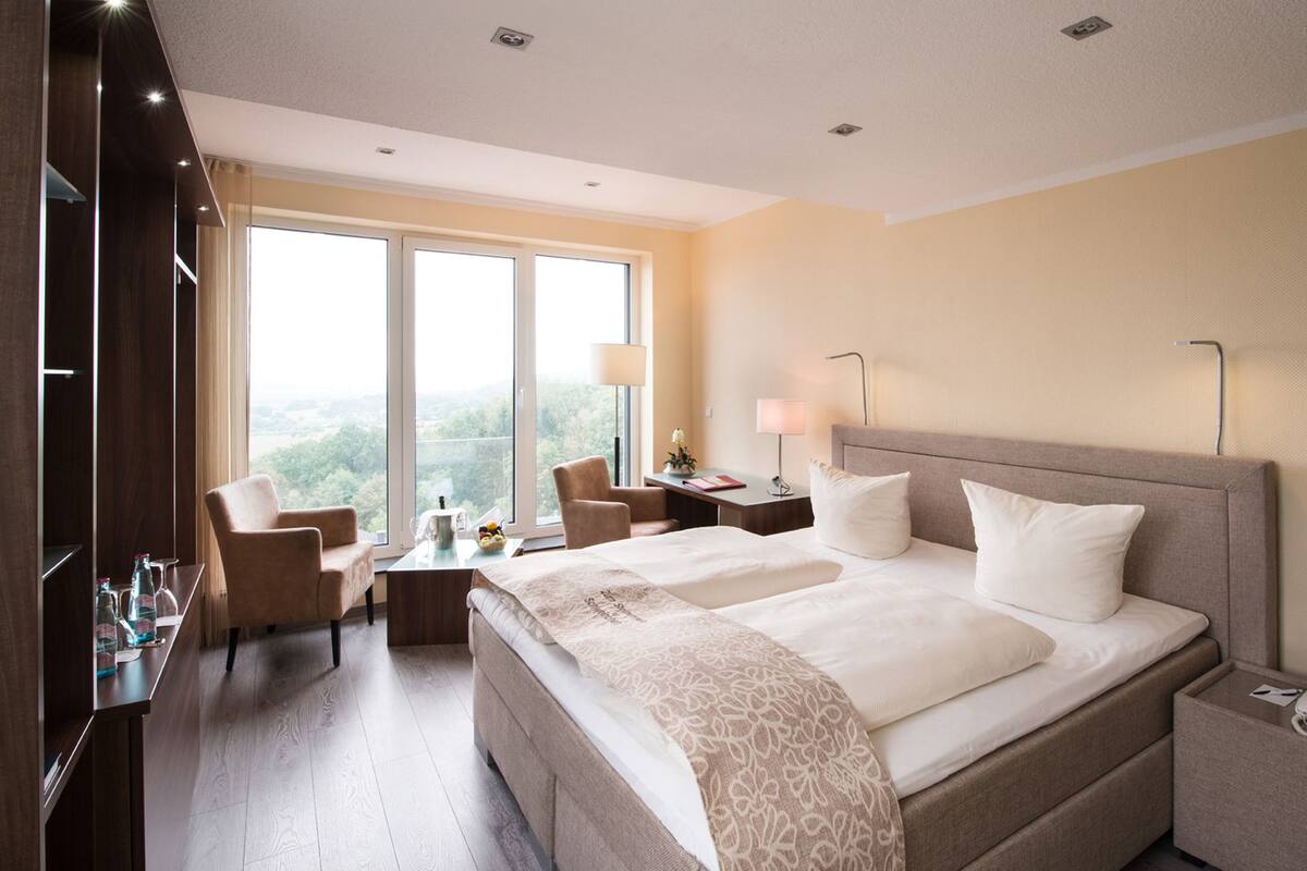 Habitación de hotel con una cama grande, dos mesitas de noche, un escritorio con una silla, un ventanal con vistas y un armario.