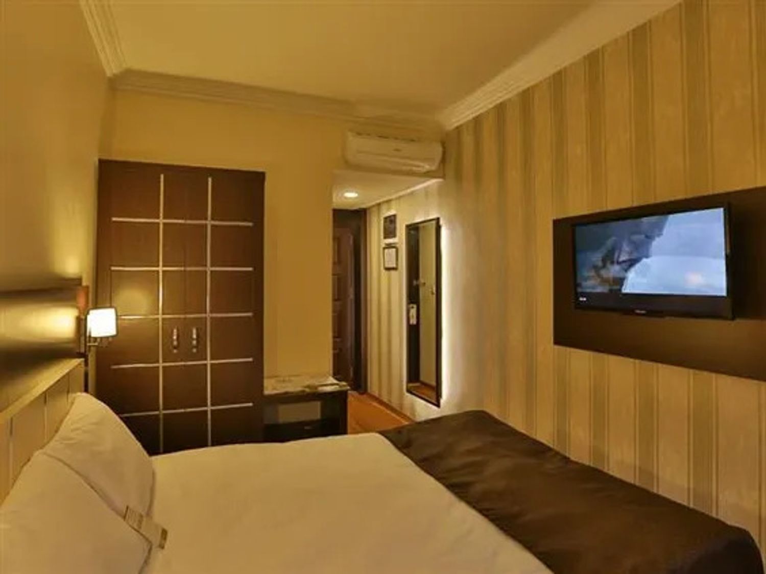 Habitación de hotel con cama, televisor montado en la pared, puerta y equipo de aire acondicionado.