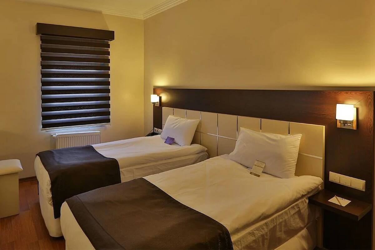 A hotel room with two beds, a window with blinds, bedside lamps, and a small table with a lamp.