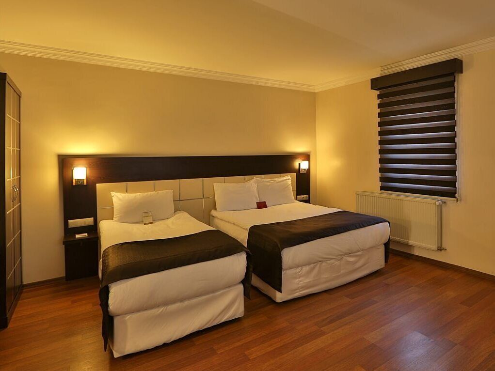 Una habitación de hotel con dos camas, piso de madera, un radiador y una ventana con persianas.