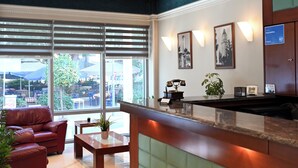 A hotel reception area with a reception desk, seating area with a sofa and chairs, large windows with a view of the outdoors, and framed pictures on the wall. - Achillion Hotel (Larissa)
