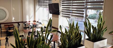 A modern indoor space with tables and chairs, large windows with blinds, and potted plants.