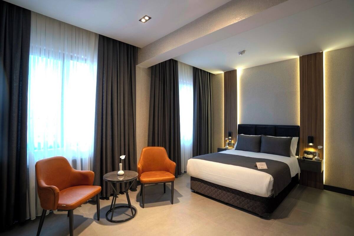 A hotel room with a large bed, two armchairs, a small table, and a nightstand with a lamp.