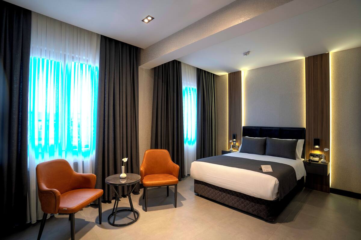 A hotel room with a large bed, two armchairs, a small table, and a nightstand with a lamp.