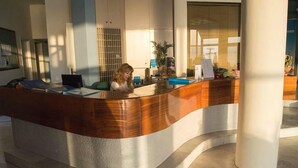 A reception desk with a curved wooden design, a seated receptionist, and a modern interior with large windows and a tiled floor. - Scaleta Beach (Rethymno)