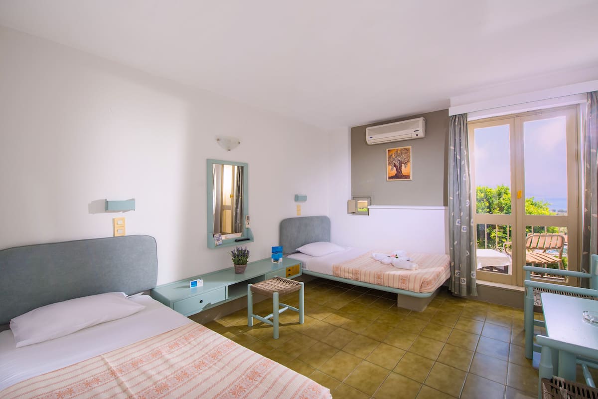 A hotel room with two beds, a small table, a chair, a window with a view, and an air conditioning unit.