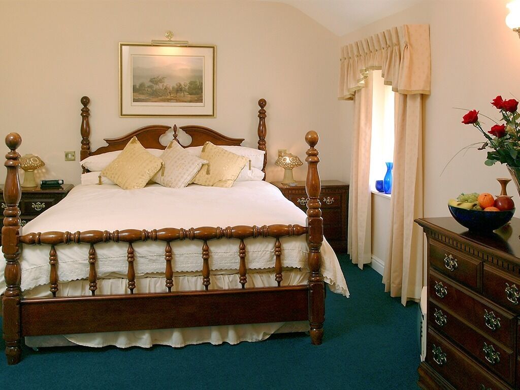 A classic four-poster bed with a ruffled skirt, a wooden nightstand with a lamp, a dresser with a fruit bowl, and a framed landscape painting above the bed.