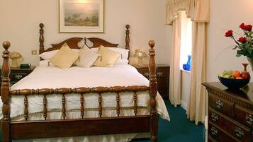 Una cama clásica con dosel y falda festoneada, una mesita de noche de madera con lámpara, un aparador con frutero, y un cuadro de paisaje enmarcado sobre la cama.