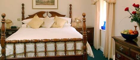 A classic four-poster bed with a ruffled skirt, a wooden nightstand with a lamp, a dresser with a fruit bowl, and a framed landscape painting above the bed.