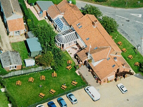 An aerial view of a residential area with a large house featuring a tiled roof, a conservatory, and a spacious lawn with outdoor furniture. - The Longham White Horse (Dereham)