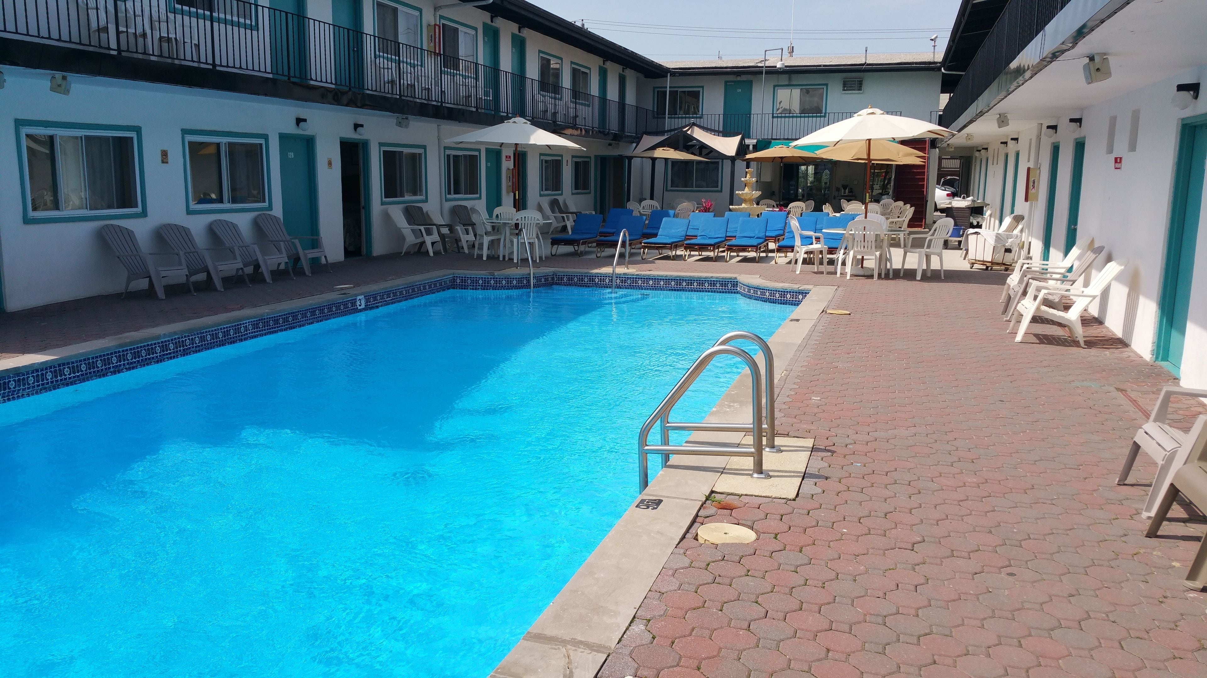 Una piscina con sillones y sombrillas, rodeada por un edificio con varias ventanas y puertas.
