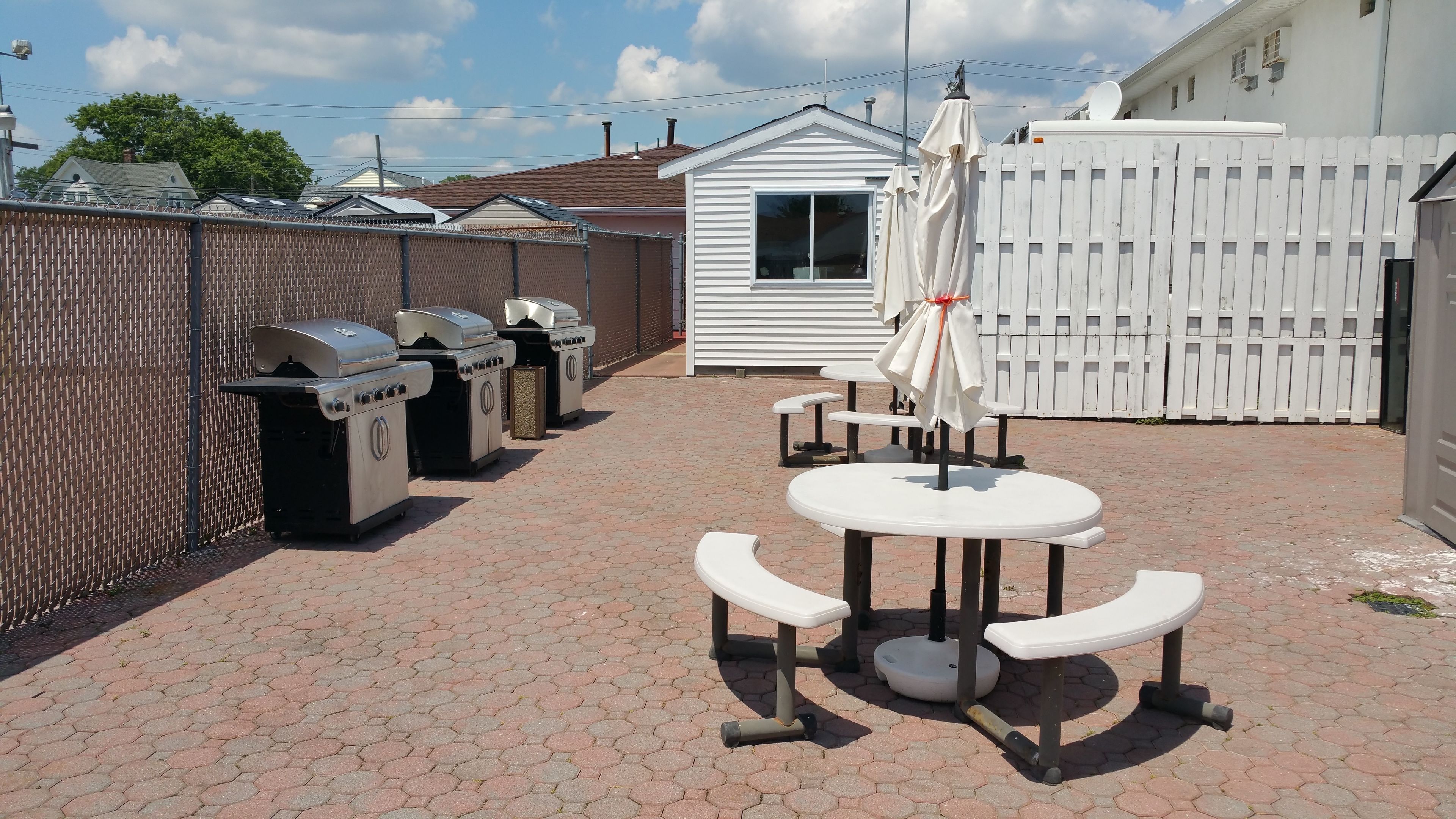 An outdoor patio with barbecue grills, a small white building, and a white fence.