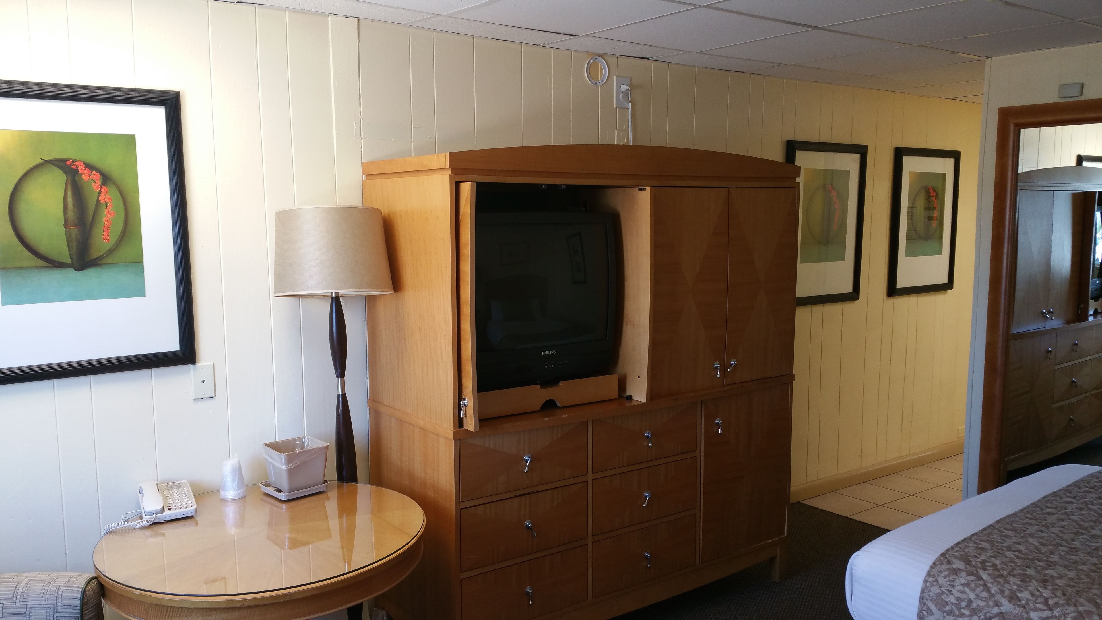 A hotel room with a wooden cabinet with a television, a round table with a lamp, and framed artwork on the walls.