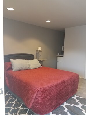 A bedroom with a red bedspread, a nightstand with a lamp, and a small desk with a mirror.