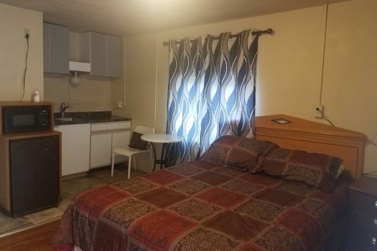 A compact room with a bed, a small kitchenette, a dining table, and a patterned curtain.