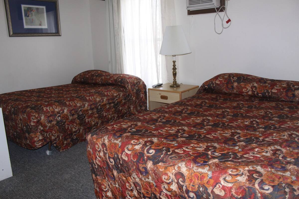 A hotel room with two beds, a bedside table with a lamp, and a framed picture on the wall.