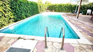 Una piscina rectangular con agua azul cristalina, rodeada por un patio de piedra y un seto.