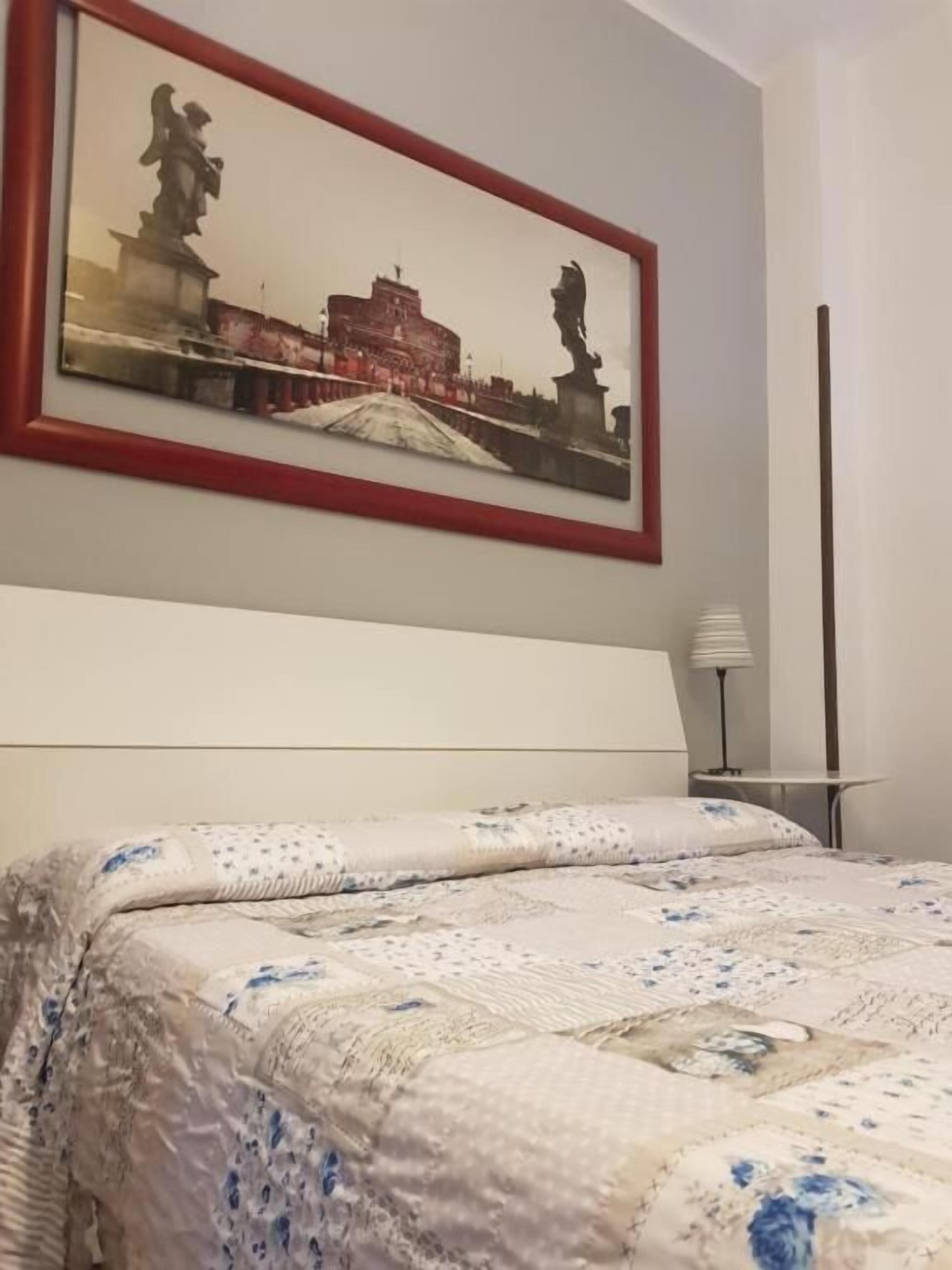 A hotel room with a bed, a framed picture on the wall, and a small table with a lamp.