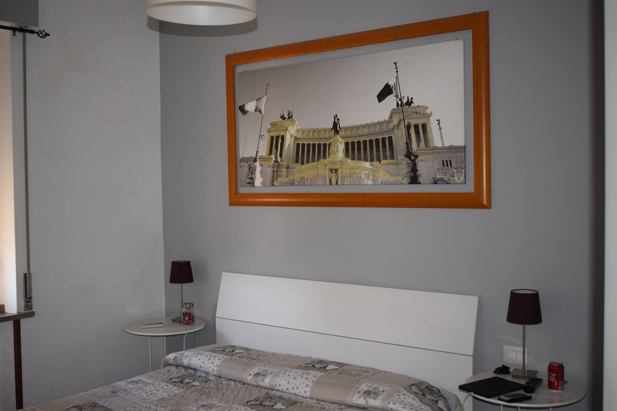 Ett hotellrum med en säng, två sängbord, en lampa på varje bord och en inramad tavla med en byggnad på väggen.