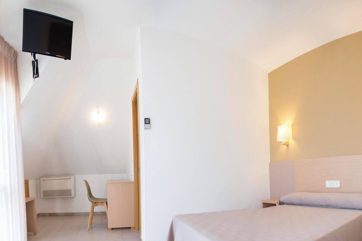 A hotel room with a bed, a desk with a chair, a TV mounted on the wall, and a bedside lamp.