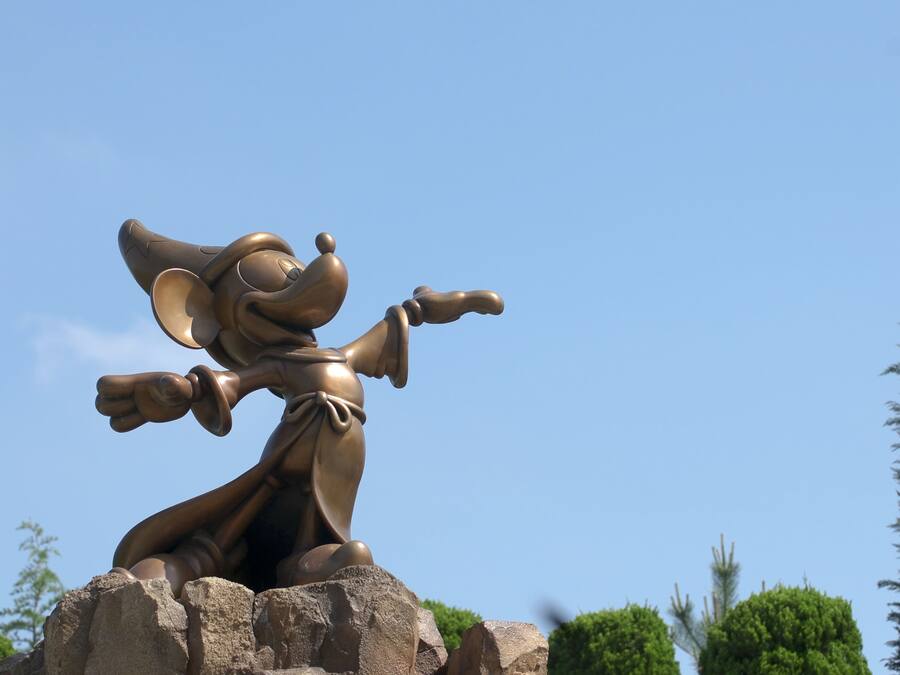 Een bronzen beeld van een personage met een hoed, armen gespreid en een wijzende gebaar, voor een helderblauwe lucht.