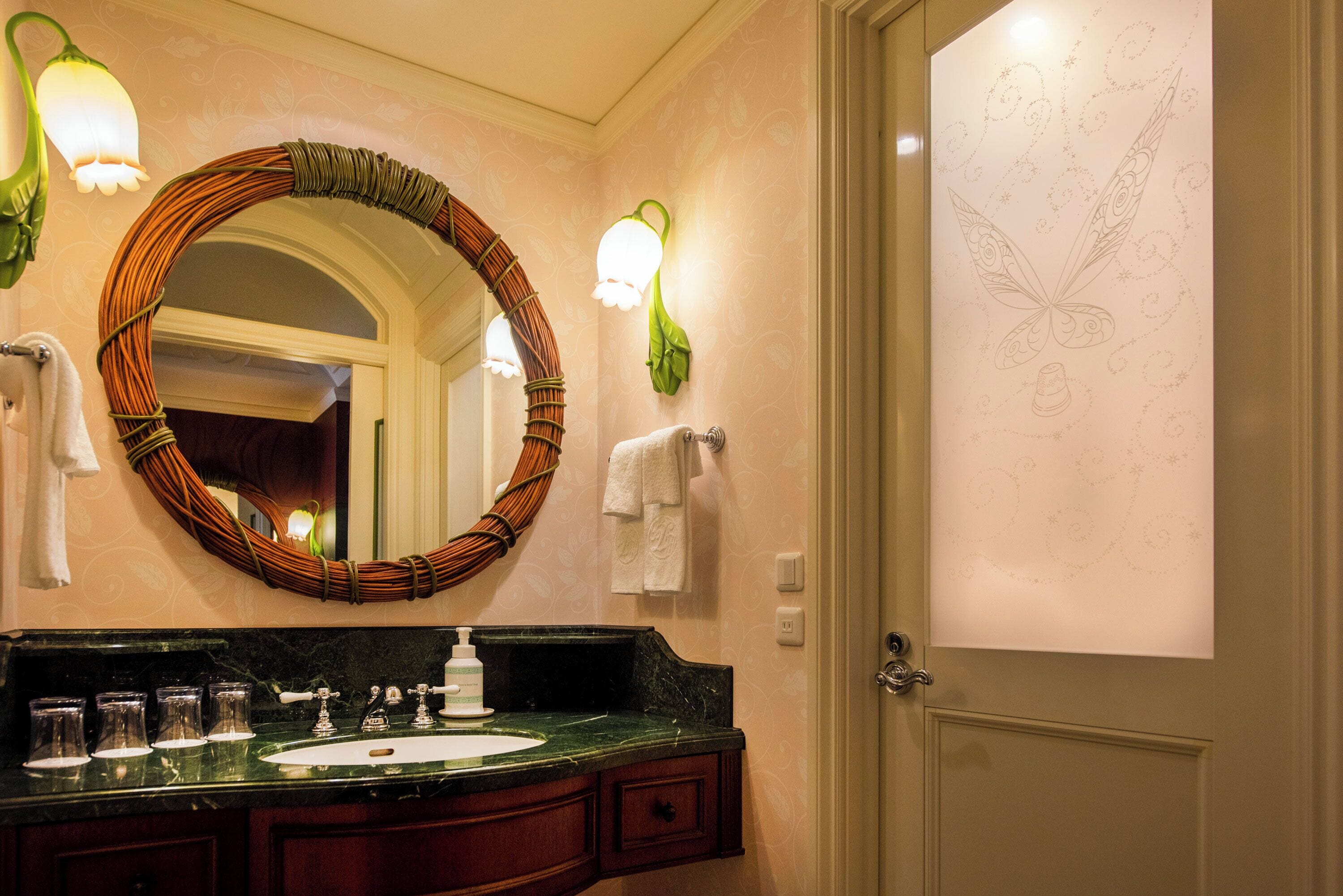 A bathroom with a round mirror, green wall sconces, a sink with multiple glasses, and a door with a butterfly design.