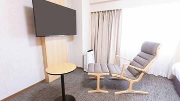 A hotel room with a flat-screen TV mounted on the wall, a round table, two cushioned chairs, a bed, and a window with curtains.