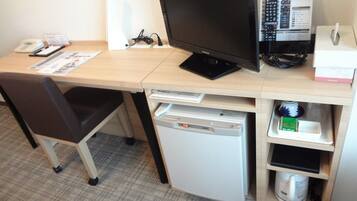 A hotel room with a desk, a computer, a small refrigerator, and a chair.