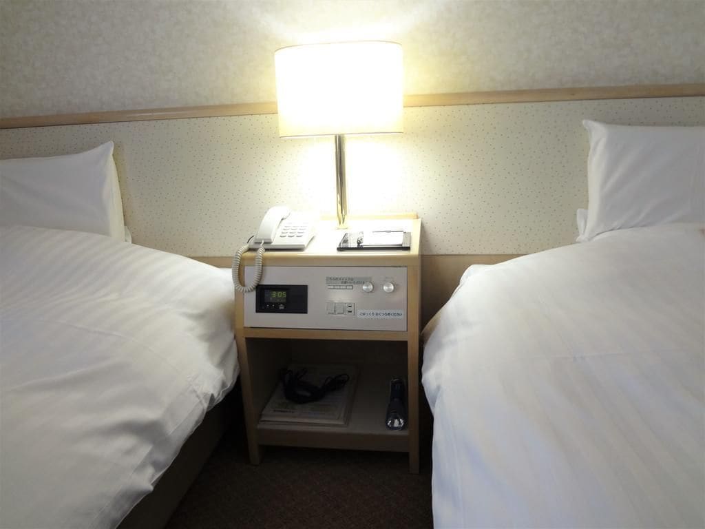 A hotel room with two beds, a bedside table with a lamp, a phone, and a digital clock.