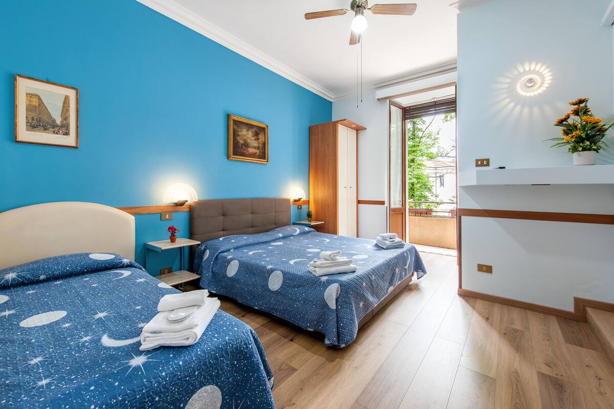 Ein Hotelzimmer mit zwei Betten, einem Deckenventilator, einem Nachttisch, einem Kleiderschrank und Blick ins Freie.