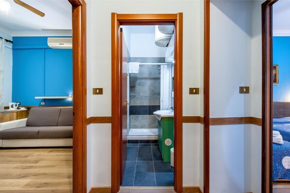 A hotel room with a wooden door leading to a bathroom, a sofa, a bed, and a small kitchenette area.