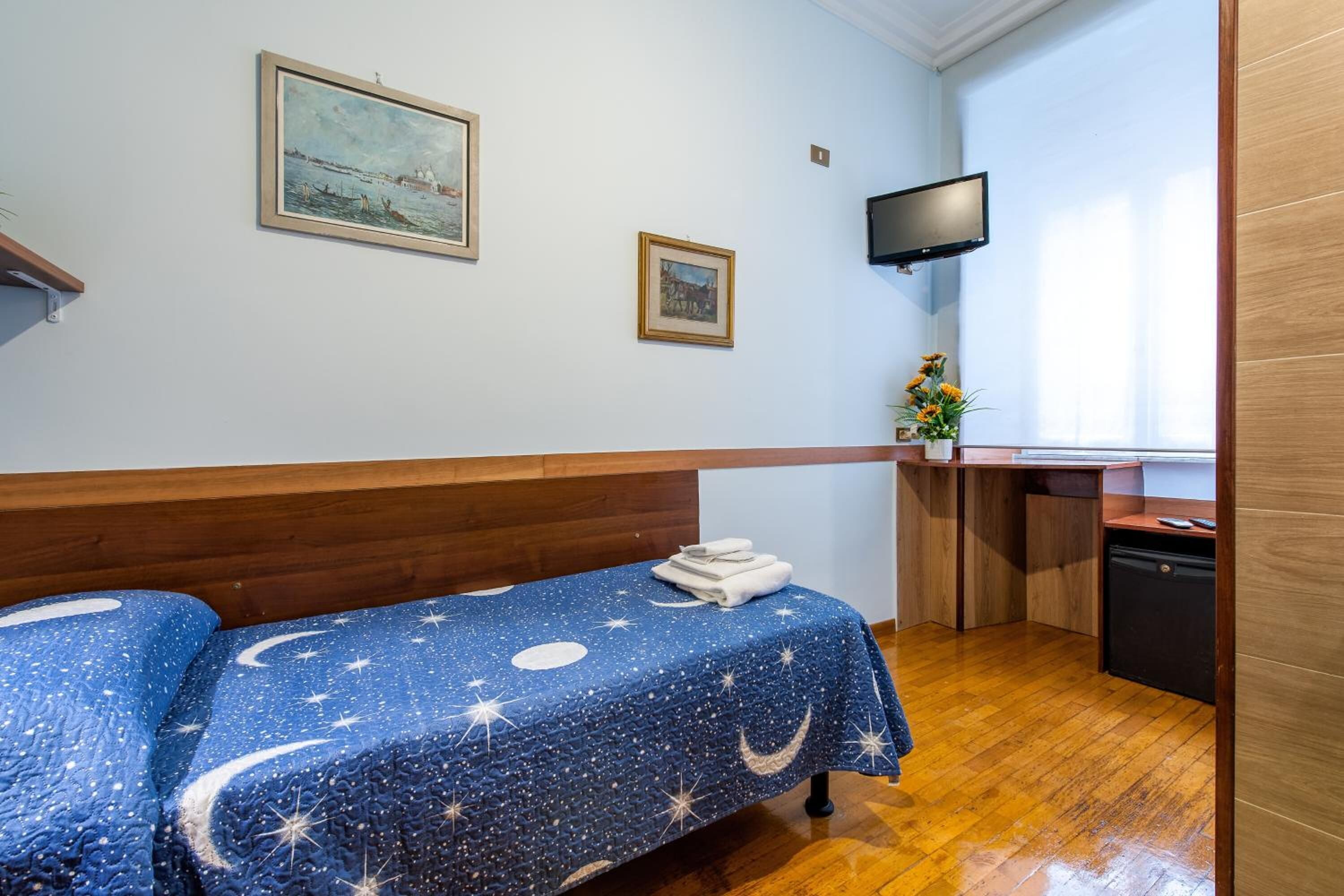A bedroom with a wooden headboard, a bed with a starry night theme, a small desk with a computer, and a TV on the wall.