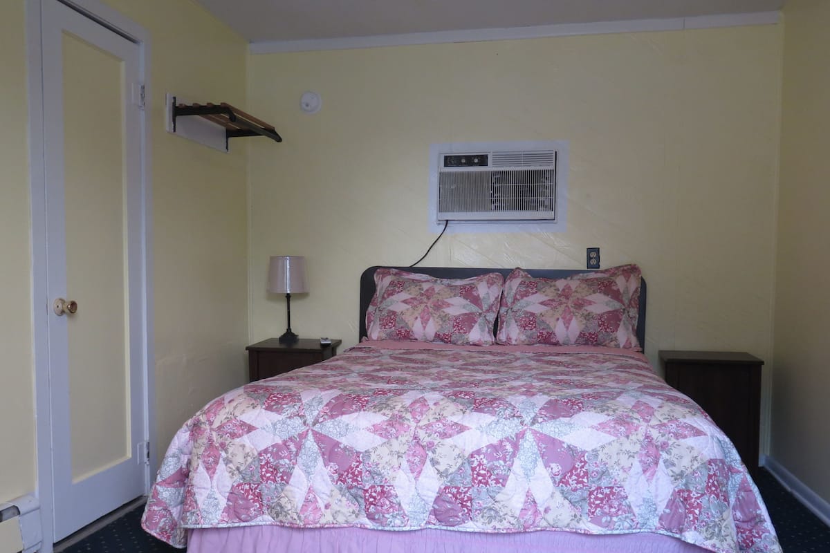 A bedroom with a bed, a nightstand, a lamp, a wall-mounted air conditioner, and a door.