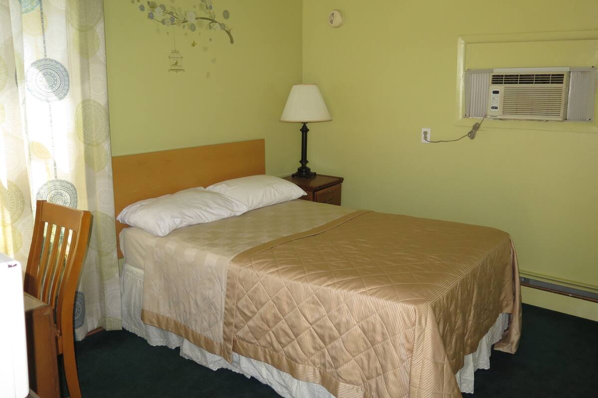 A neatly made bed with a quilted cover, a bedside lamp, and a wall-mounted air conditioning unit.