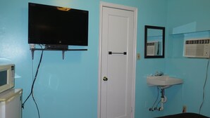 A room with a television mounted on the wall, a white door, a sink with a mirror above it, and a heater on the floor. - Pillow Talk Motel (Wellington)