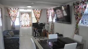 A living room with a sofa, a dining table with chairs, a TV mounted on the wall, and curtains with a geometric pattern. - Princess Apartment (Mangrove)