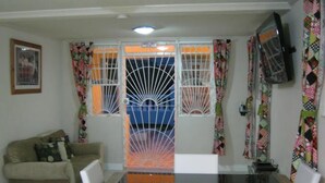 A living room with a beige sofa, a glass-top dining table, and a TV mounted on the wall. - Princess Apartment (Mangrove)