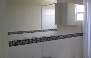 Une salle de bain avec des carreaux blancs, un pare-baignoire carrelé, un miroir et une fenêtre avec des rideaux.