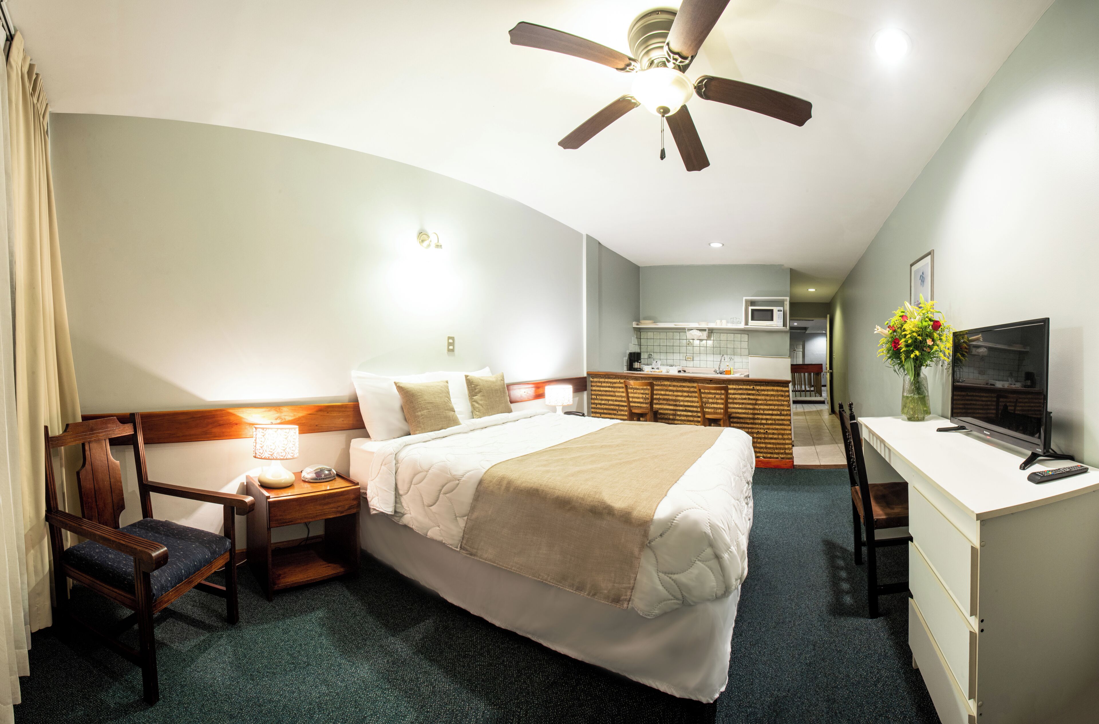 Quarto de hotel com cama, escrivaninha, cadeira, mesa de cabeceira, abajur, televisão e ventoinha no teto.