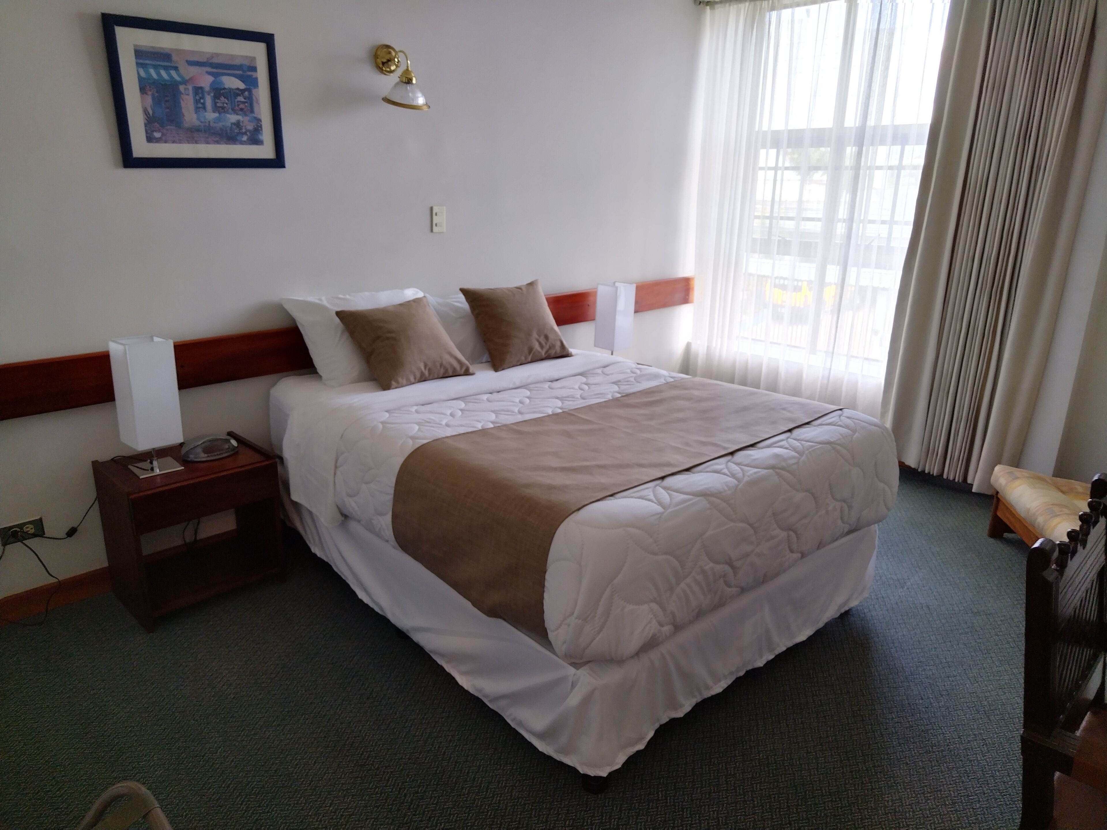 Una cama bien hecha con una colcha blanca y marrón, una lámpara de noche y un cuadro en la pared.