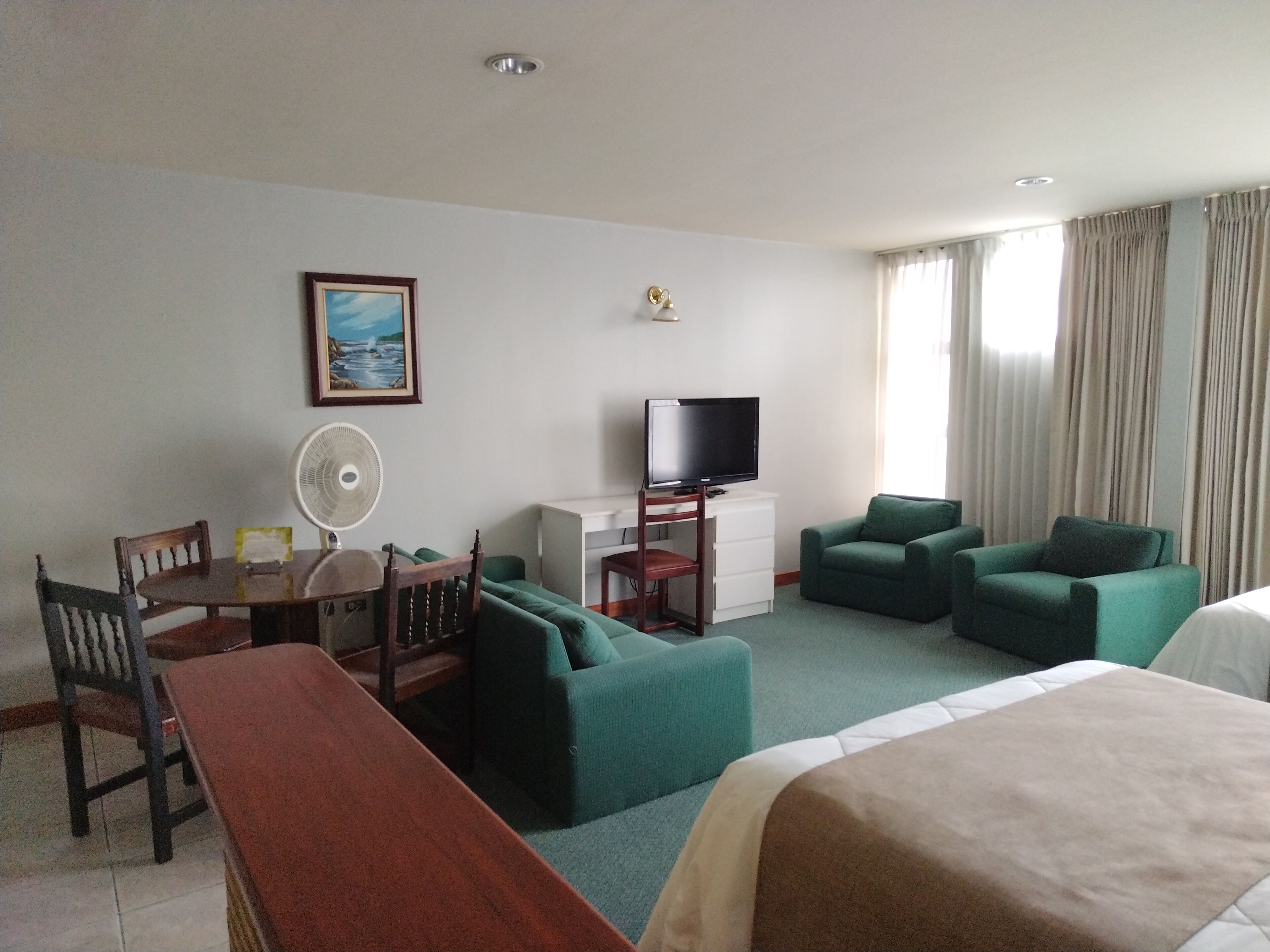 Una habitación de hotel con una cama, un escritorio con una silla, un televisor y dos sillones verdes.