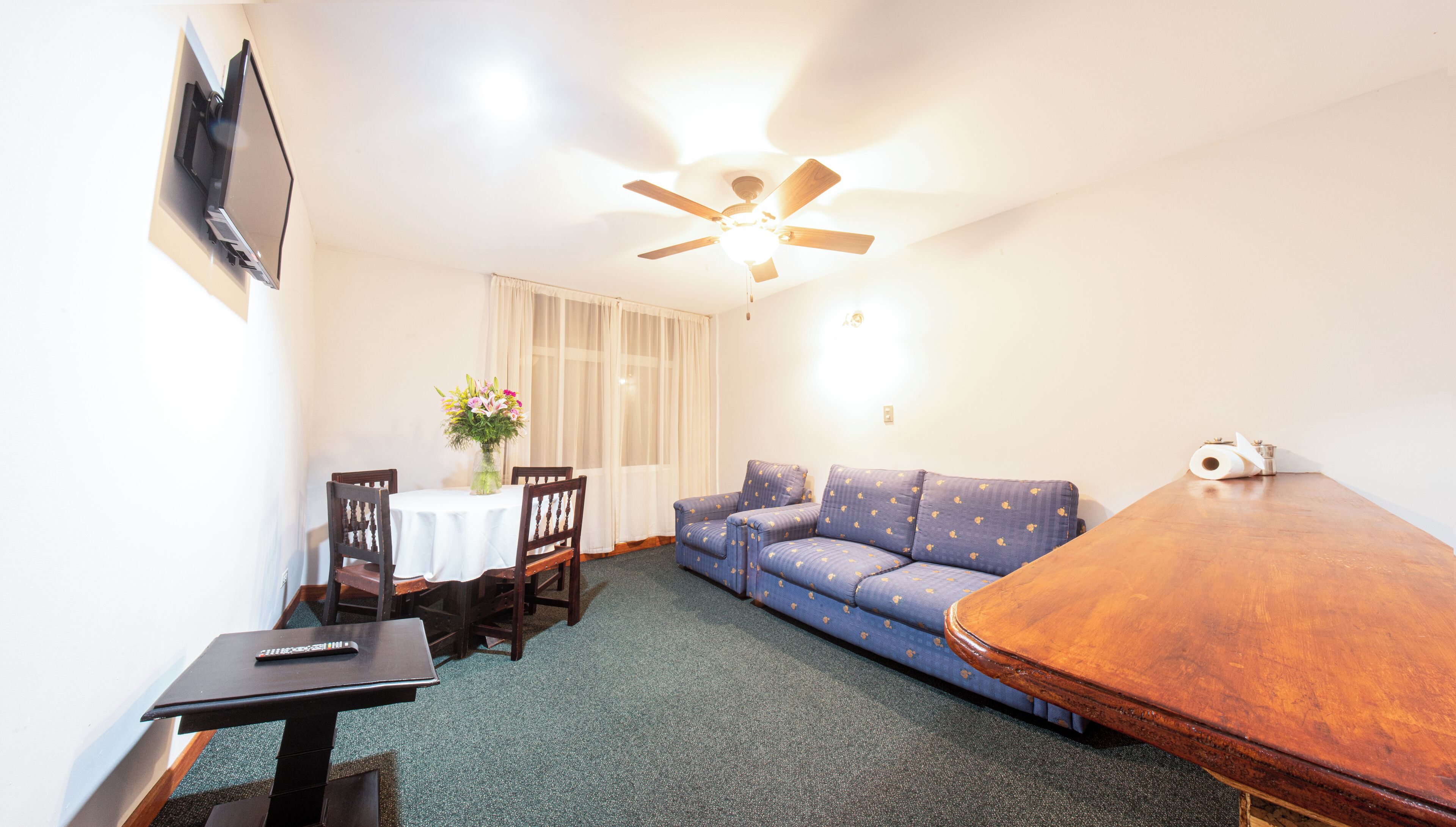 A room with a wooden table, a sofa, a TV mounted on the wall, and a ceiling fan.