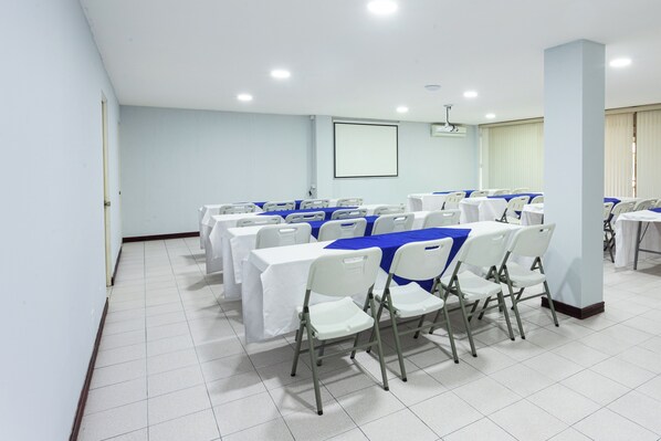 Uma sala de conferência com cadeiras e mesas brancas, uma tela de projetor e um quadro branco.