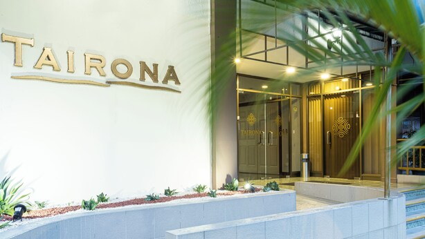 The exterior of a hotel named 'TAIRONA' with a modern entrance, glass doors, and a well-maintained garden area.