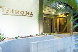 Exterior de um hotel chamado 'TAIRONA', com entrada moderna, portas de vidro e uma área de jardim bem cuidada.