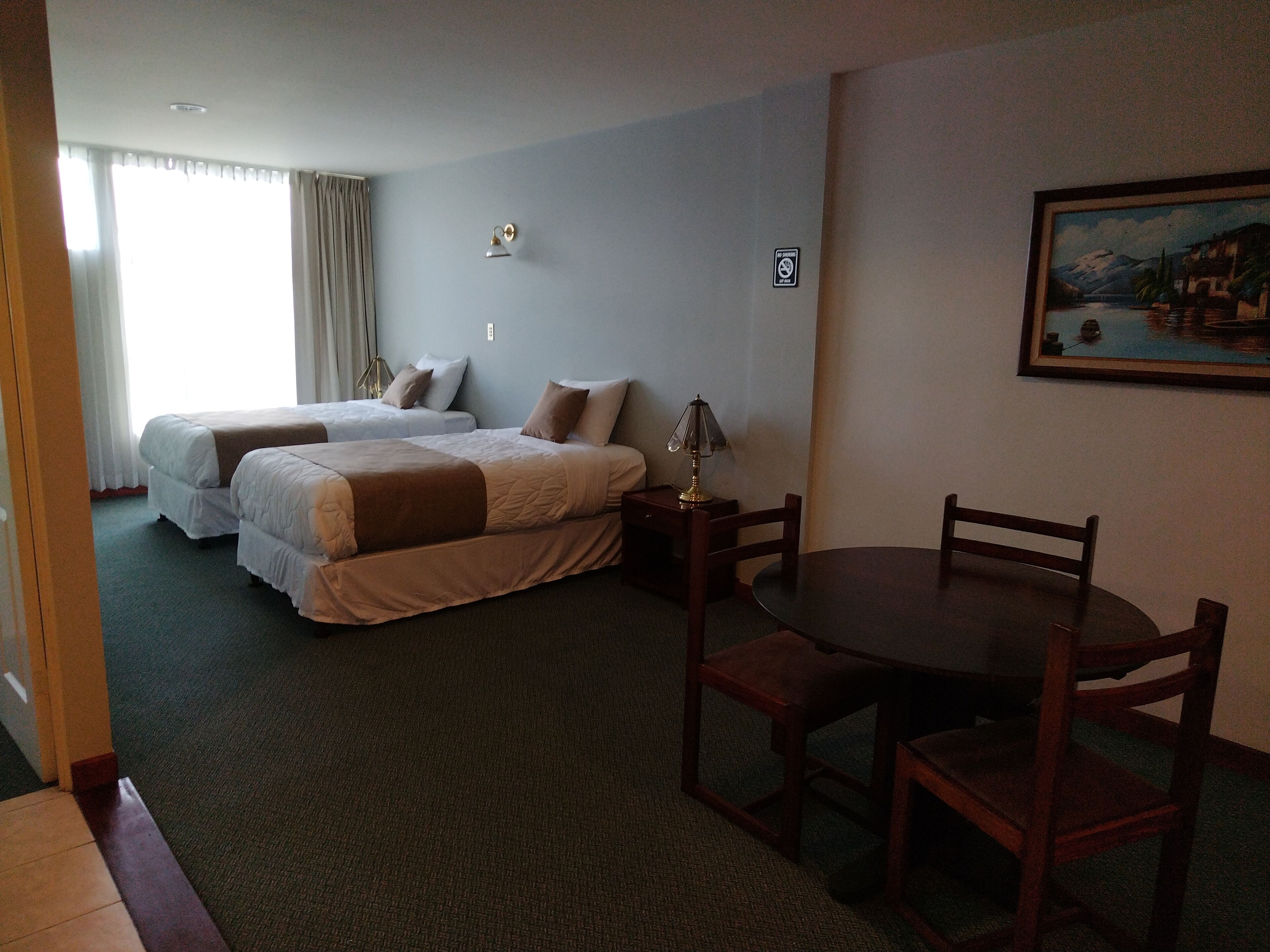 A hotel room with two beds, a dining table, and a painting on the wall.