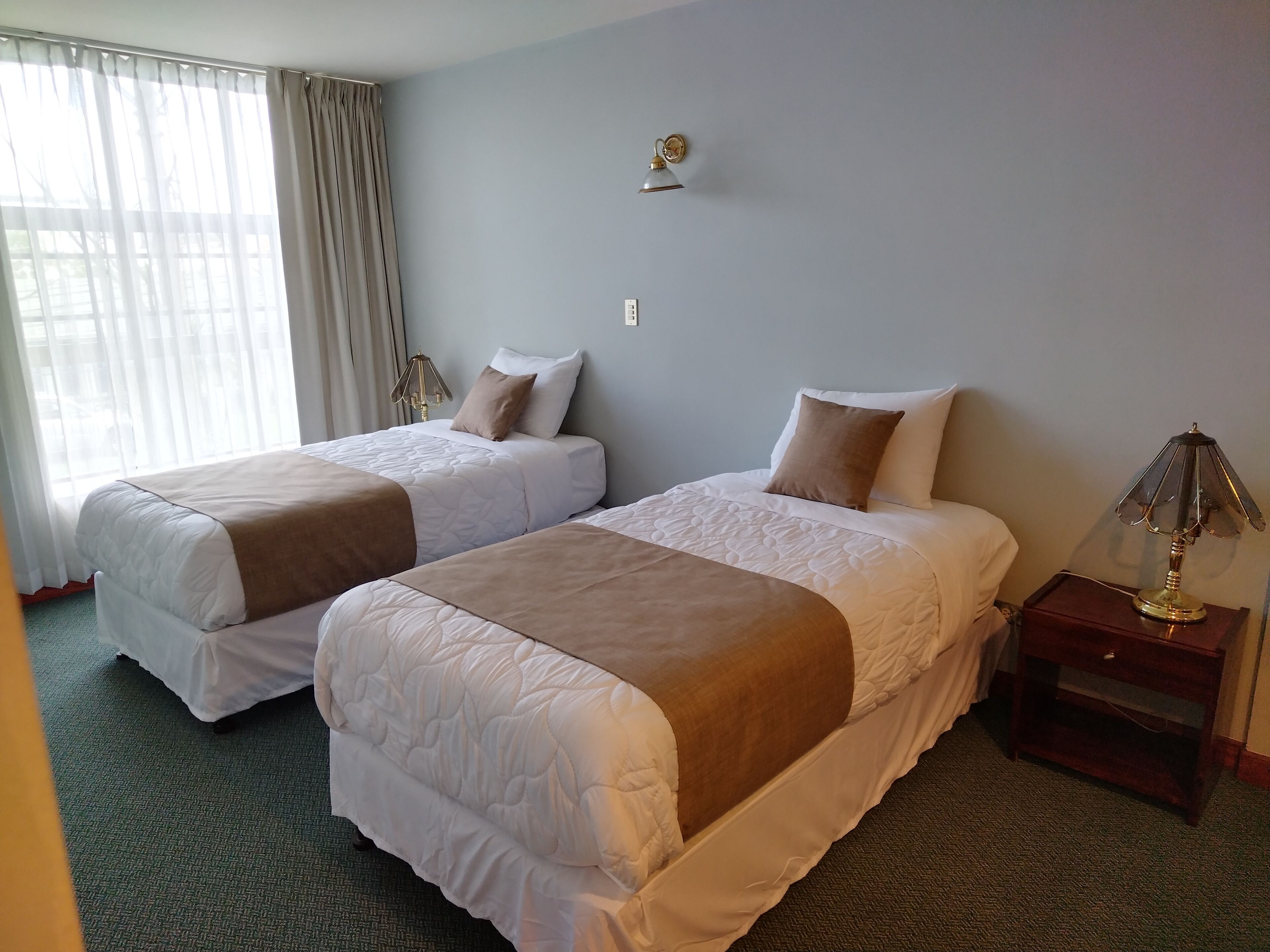 A hotel room with two beds, a nightstand, a lamp, and a window with curtains.