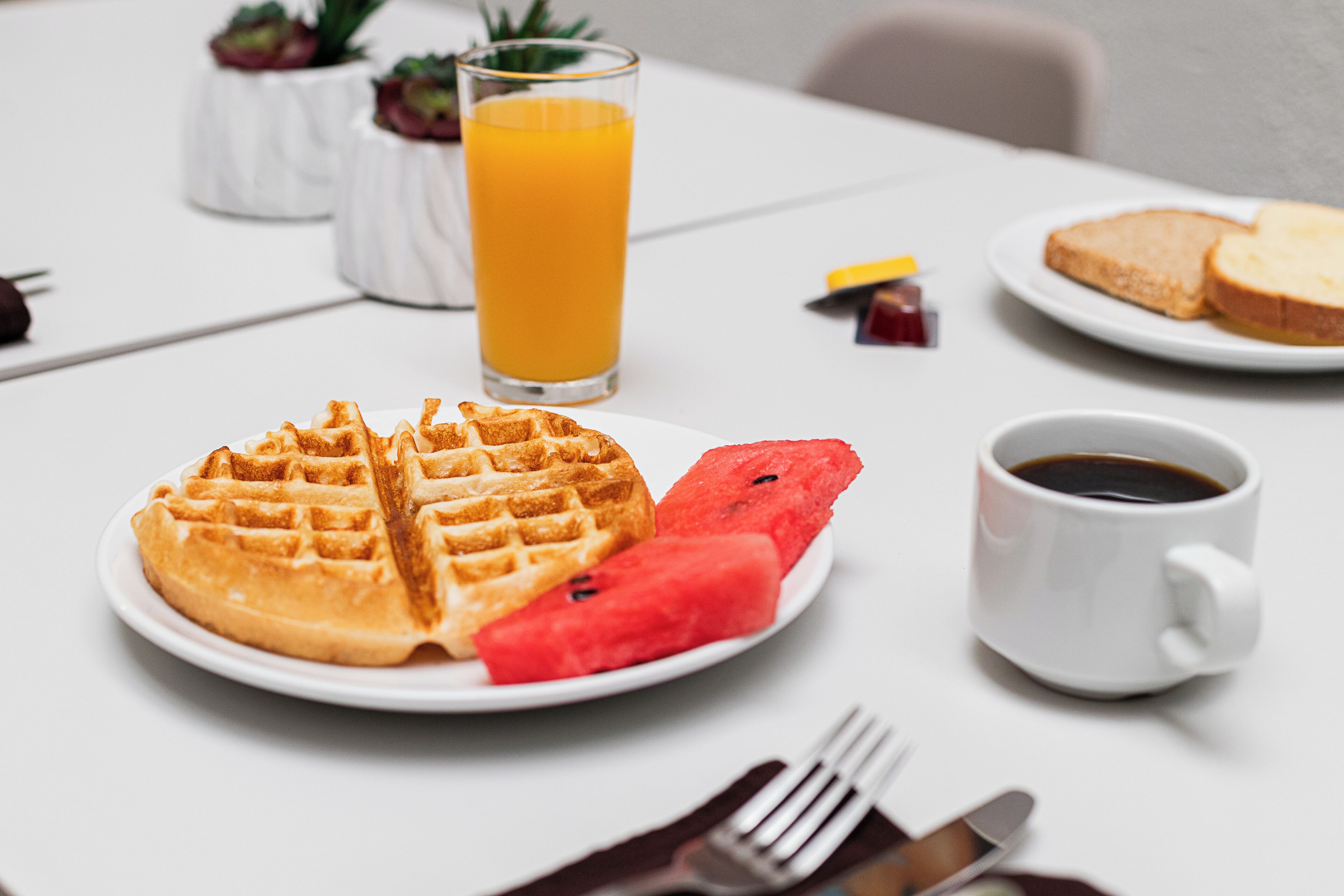 A breakfast setting with waffles, watermelon, orange juice, coffee, and toast on a white table.