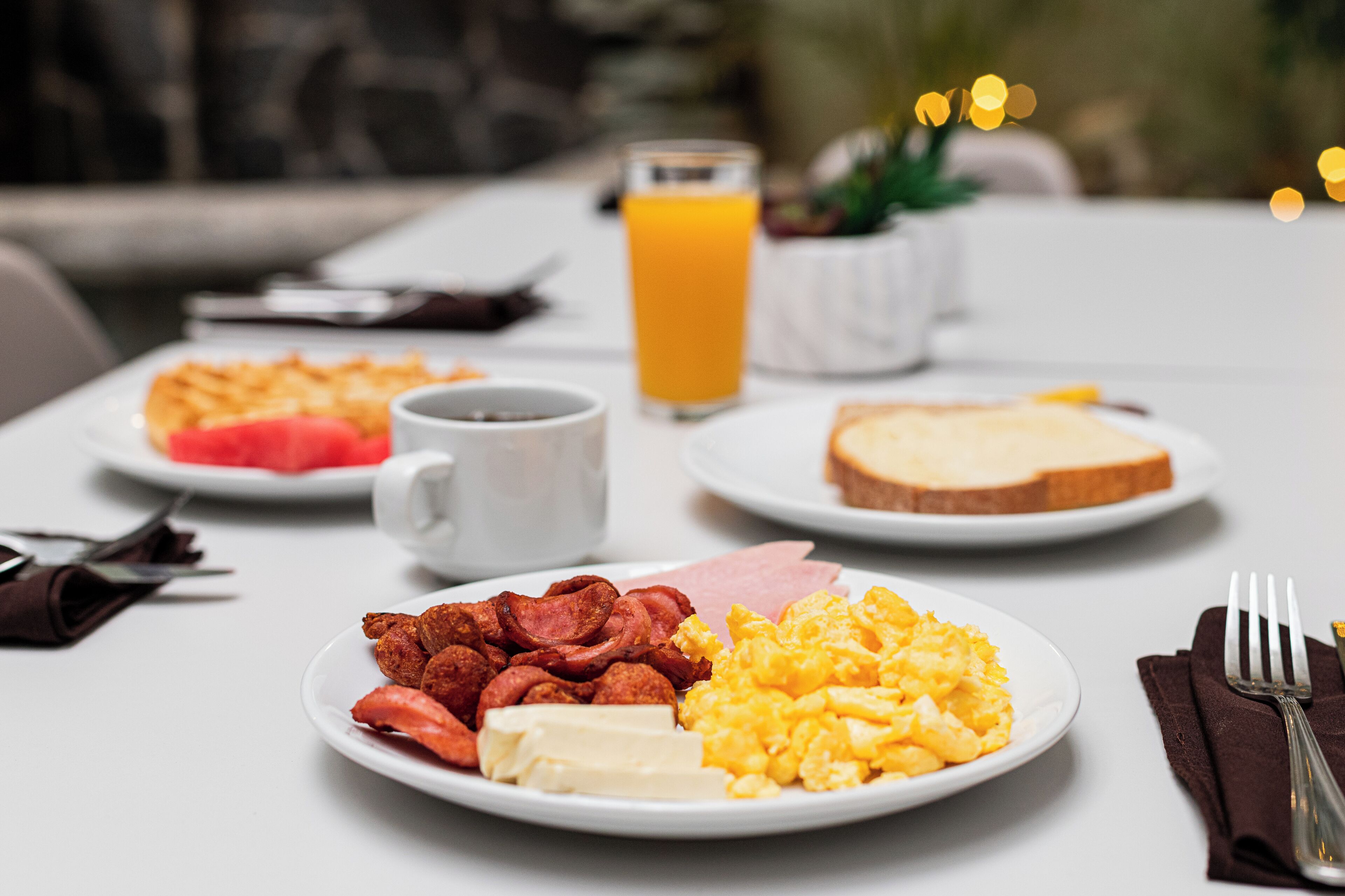 Ett frukostbord med tallrikar fyllda med ägg, bacon, skinka, rostat bröd och ett glas apelsinjuice.